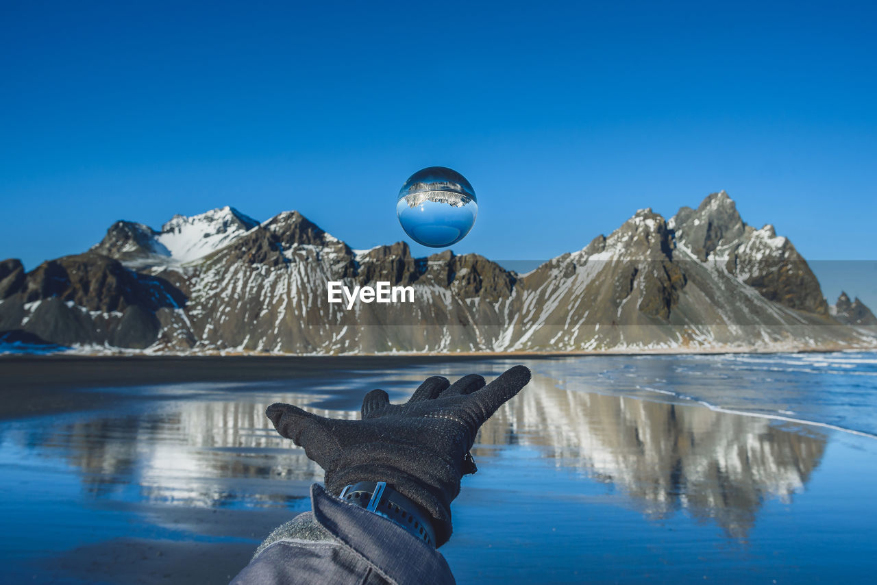 Cropped hand wearing glove while levitating crystal ball by lake against mountains during winter
