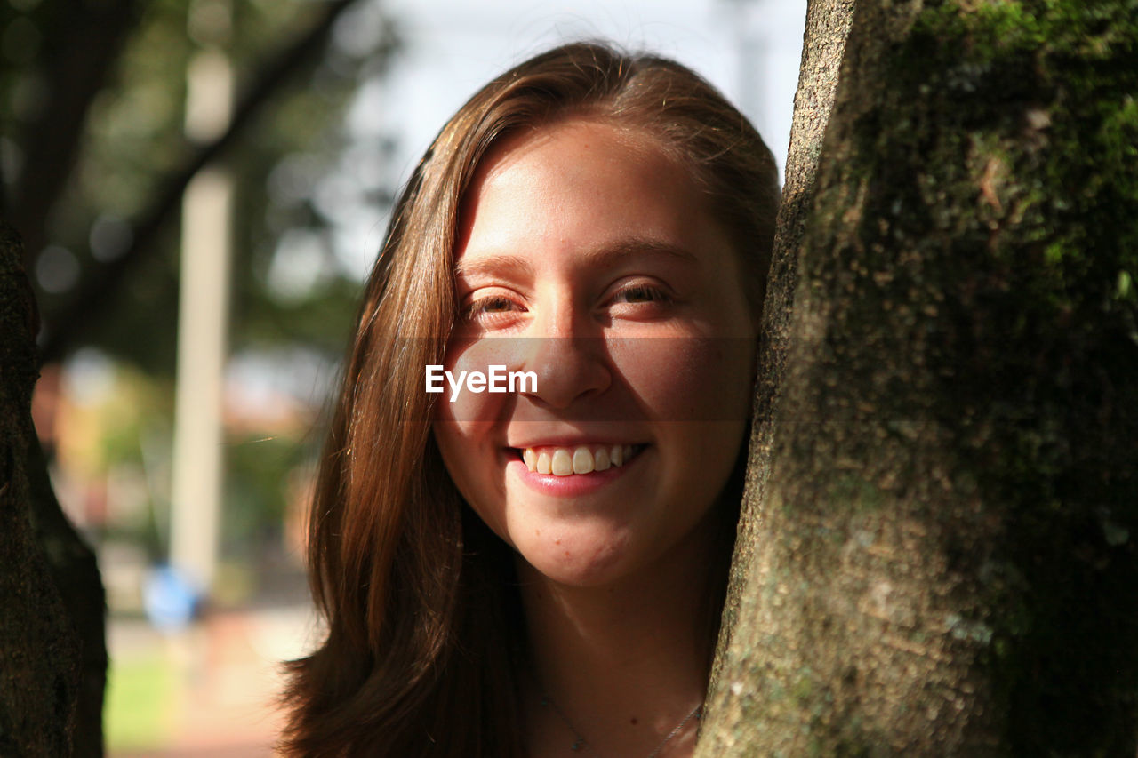 Close-up portrait of woman smiling while standing behind tree trunk