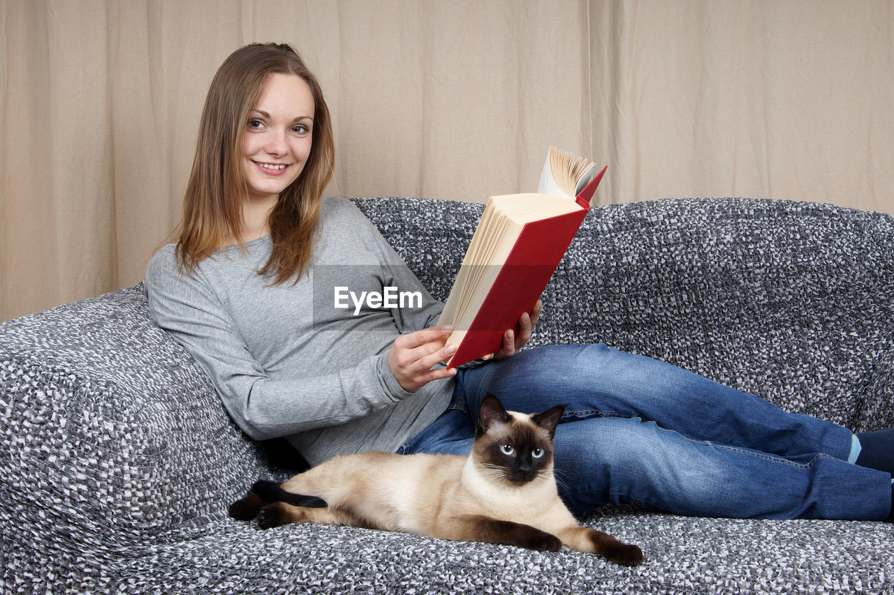 Portrait of beautiful woman with siamese cat on couch at home