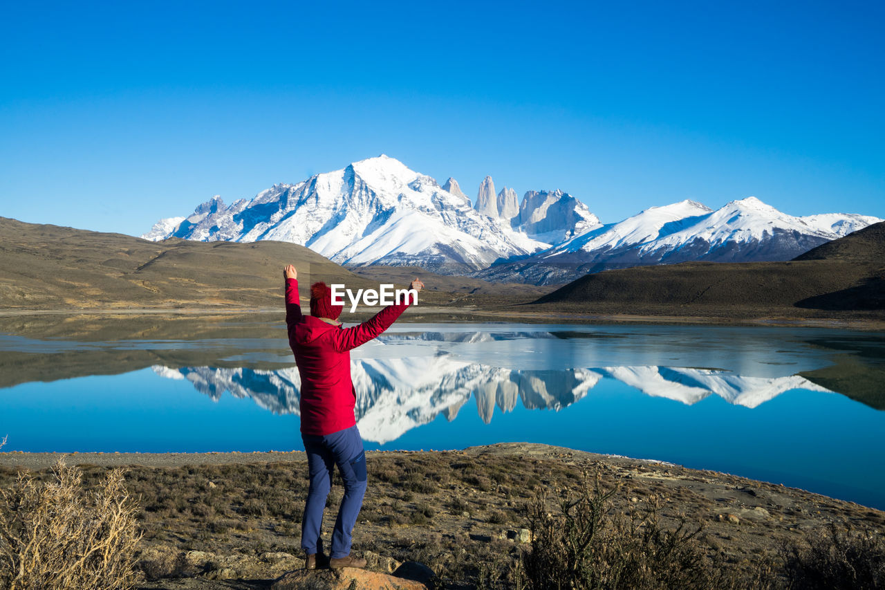 Rear view of man with arms raised standing at lake against blue sky