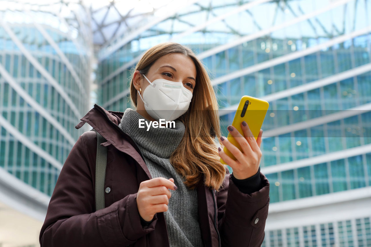 Business woman covering face with protective mask holding mobile phone with modern city background