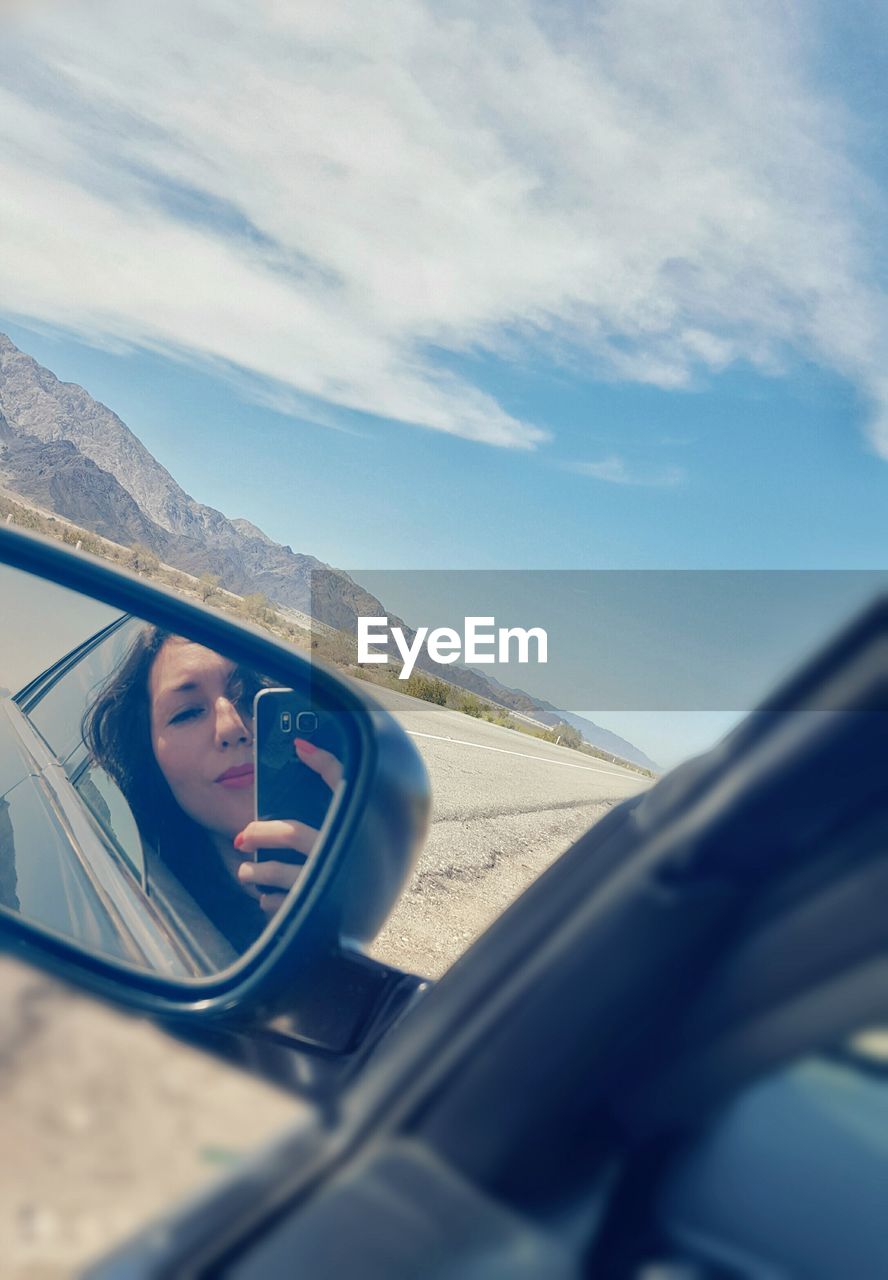 Young woman with mobile phone reflecting on side-view mirror