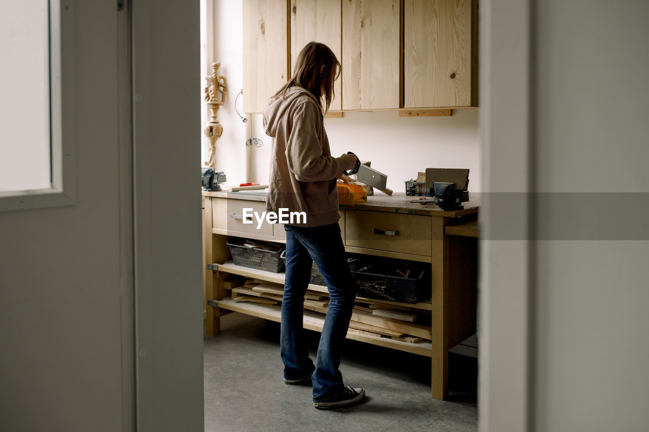 Female teenage student cutting wood during carpentry class seen through doorway