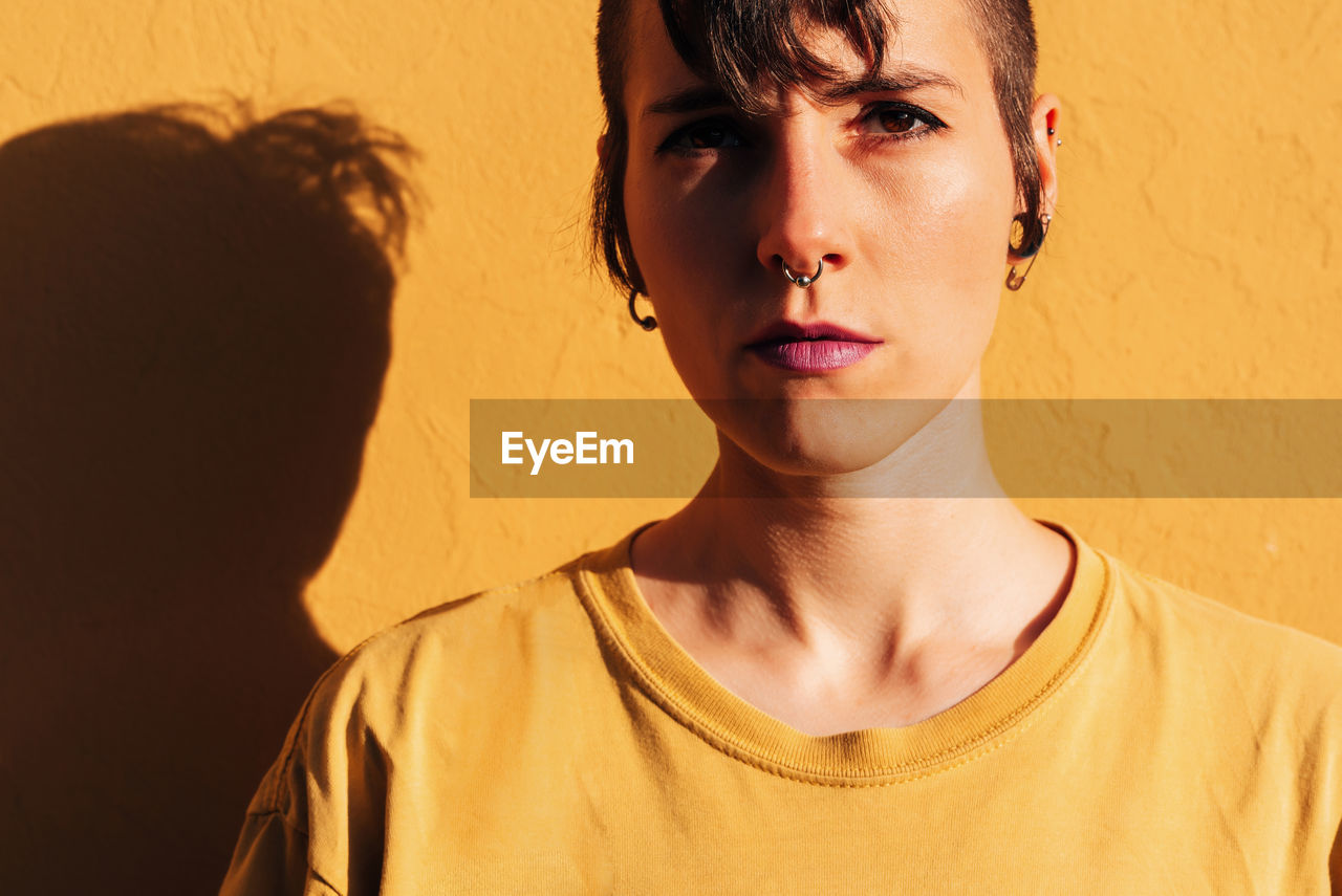 Modern woman with stylish haircut and piercing while standing near yellow wall on sunny day on street