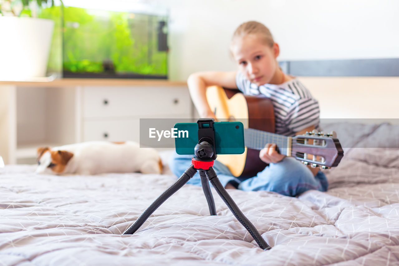 A child plays an guitar at home, looking into a smartphone on a tripod. 