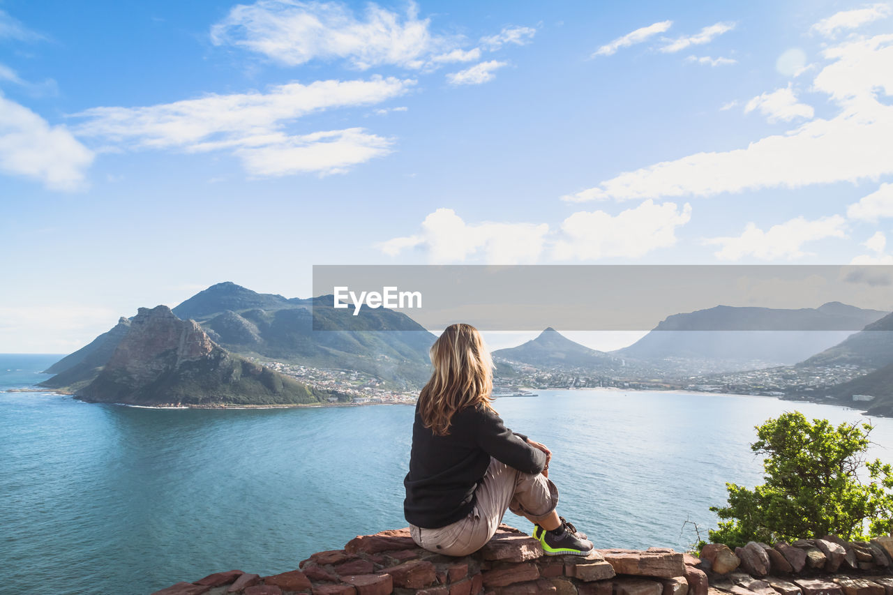 Side view of woman sitting on stone retaining wall while looking at hout bay against sky