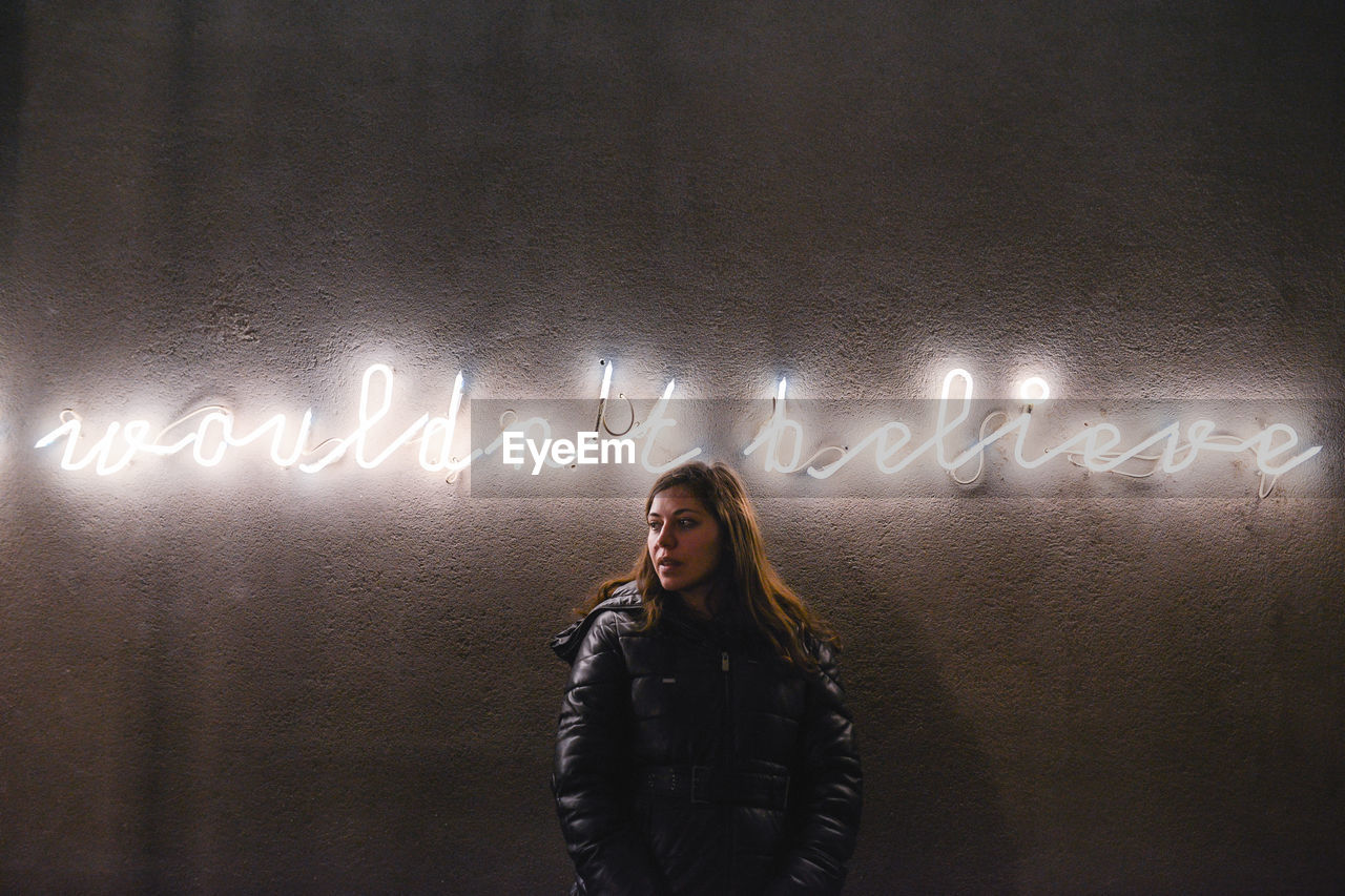 Woman looking away standing by illuminated text on wall