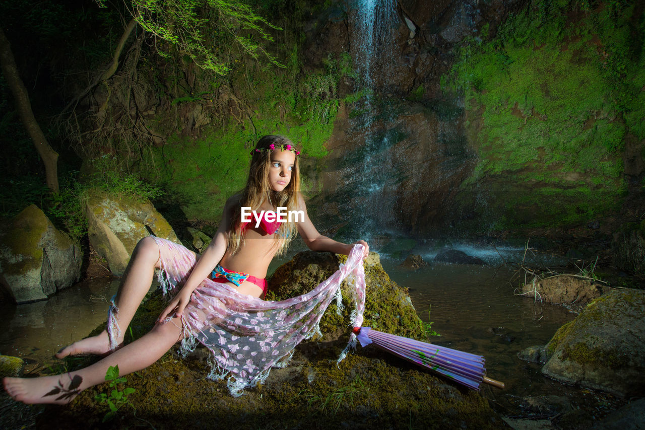 Full length of girl wearing bikini posing on rock against waterfall