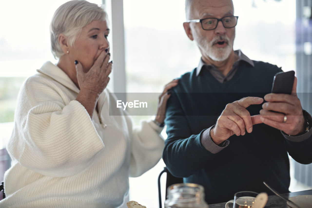 Shocked senior couple using smart phone while sitting in living room