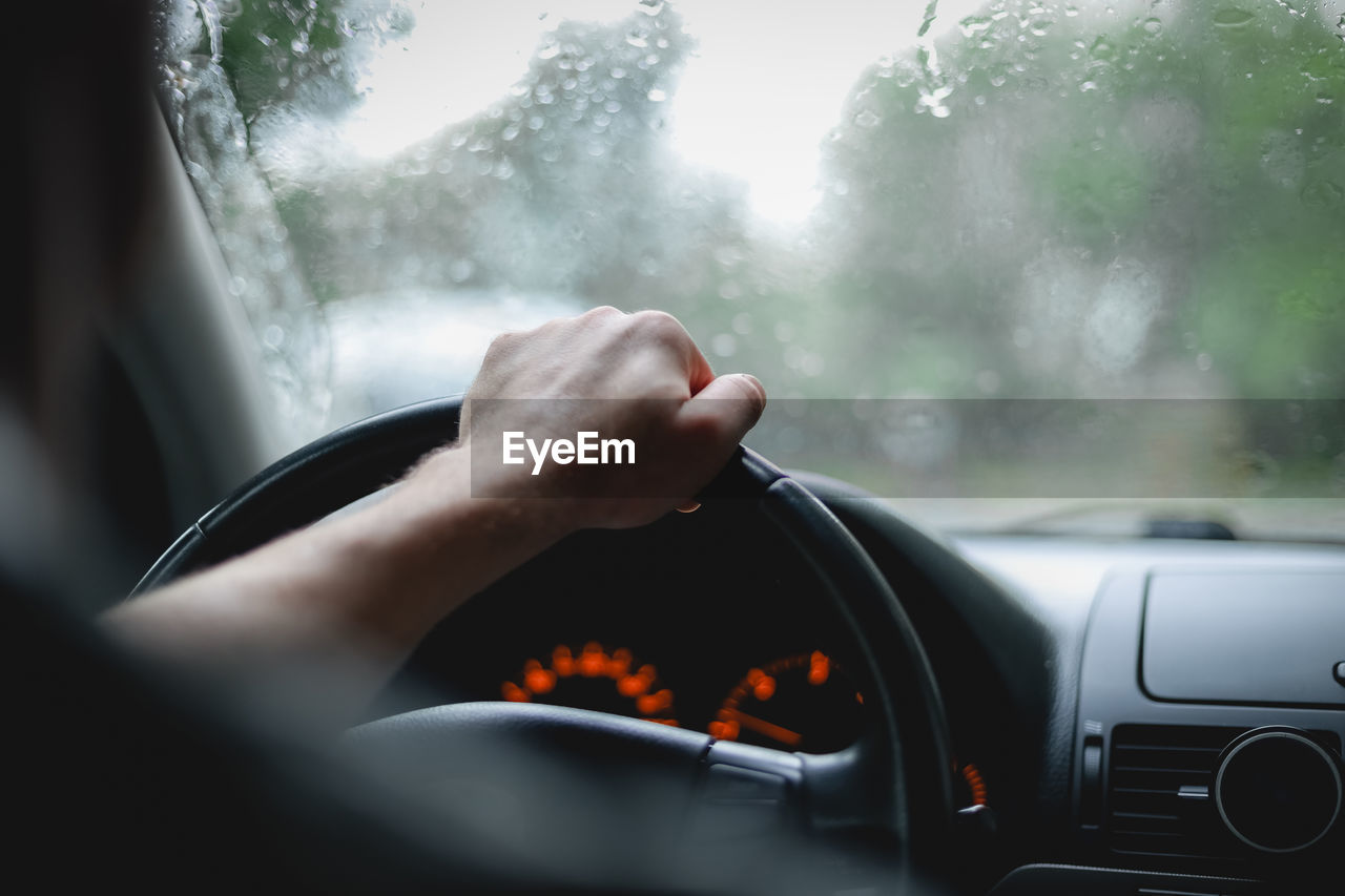 Young caucasian guy keeps his hand on the steering wheel while driving a car on a rainy day