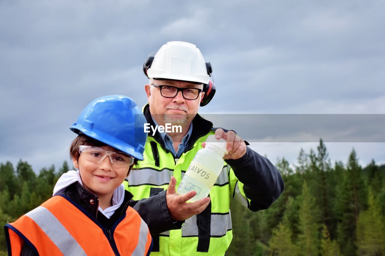 Portrait of smiling grandfather and grandson wearing hardhats against cloudy sky