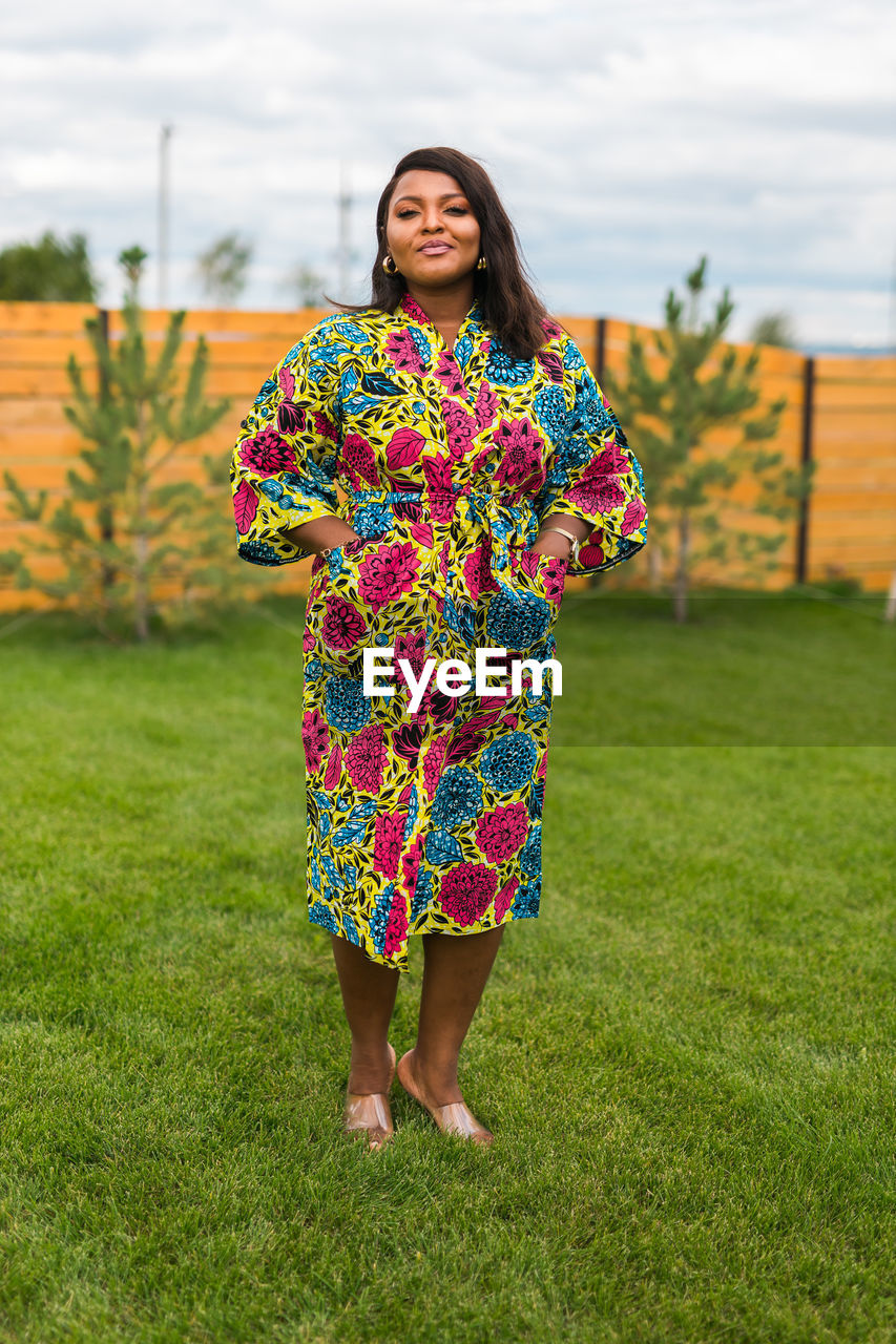 one person, spring, smiling, dress, flower, women, grass, happiness, full length, adult, yellow, plant, portrait, standing, nature, clothing, front view, looking at camera, young adult, emotion, fashion, green, cheerful, floral pattern, lifestyles, multi colored, sky, pattern, leisure activity, outdoors, cloud, female, day, hairstyle, architecture, field, flowering plant, enjoyment, smile, person, casual clothing, teeth, brown hair, traditional clothing, holding