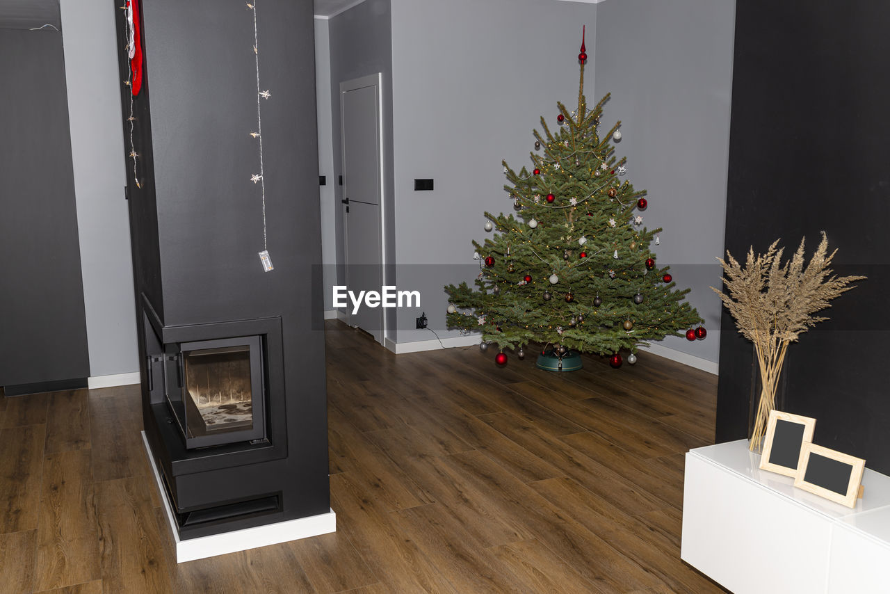 A spruce christmas tree decorated with baubles standing in the hall of a modern house, fireplace