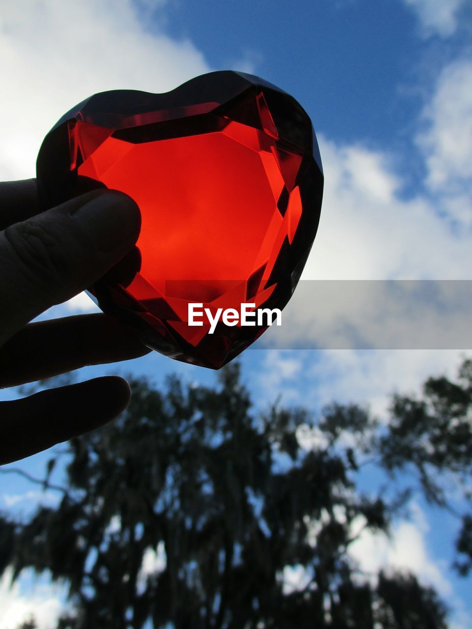 Cropped image of hand holding heart shaped red crystal against sky