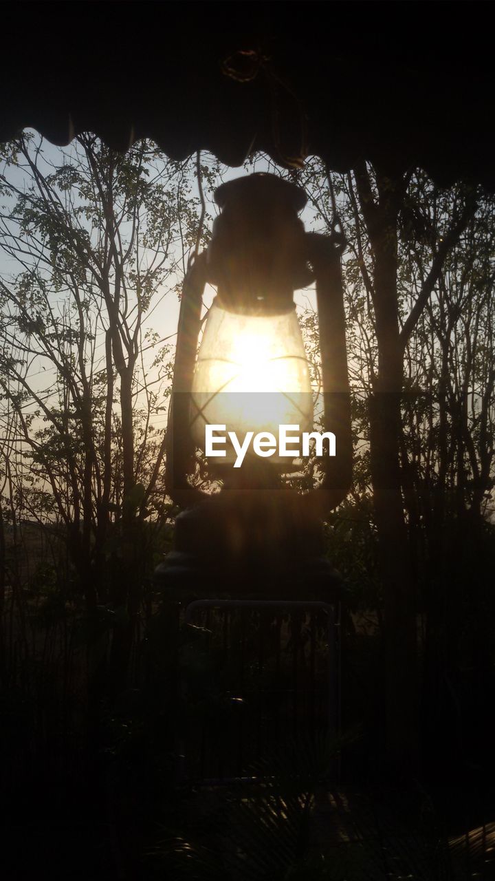 Low angle view of oil lantern hanging from roof against bare trees during sunset