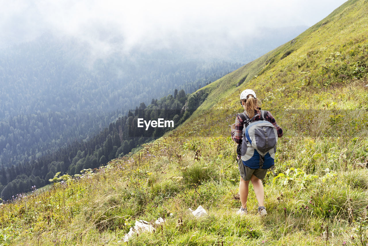 Young woman in cap and plaid shirt with big backpack hiking in green mountains against fog in summer 