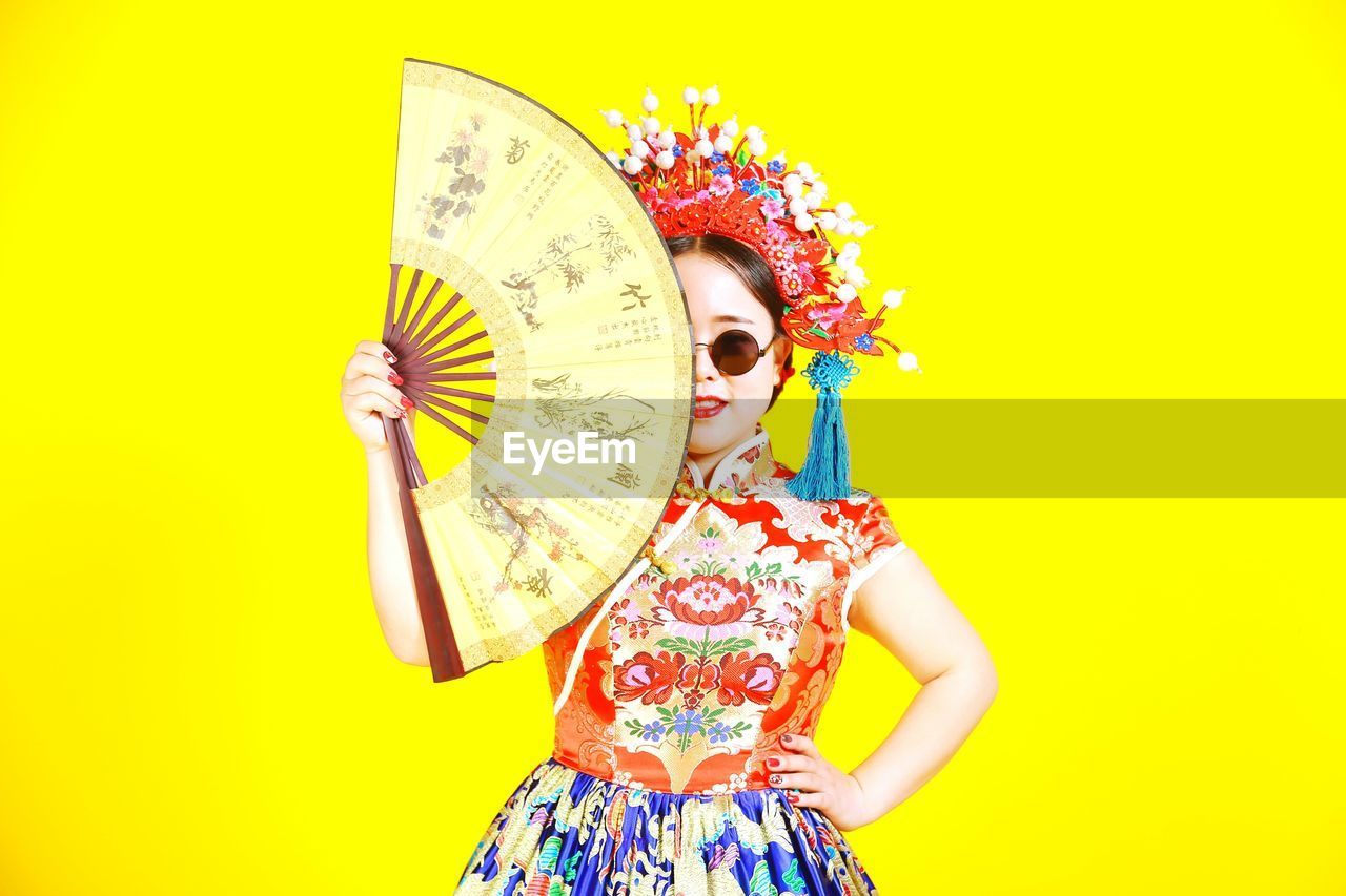 Woman in traditional clothing holding hand fan while standing against yellow background