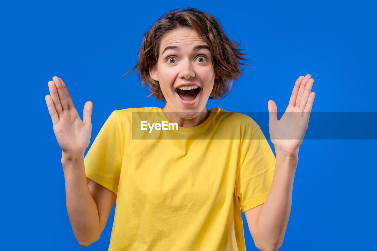 blue, emotion, shouting, happiness, yellow, one person, excitement, finger, positive emotion, studio shot, portrait, gesturing, mouth open, adult, person, smiling, colored background, cheerful, casual clothing, waist up, women, fun, looking at camera, joy, sky, facial expression, teenager, sign language, blue background, hand, young adult, arm, human mouth, front view, vitality, limb, copy space, ecstatic, child, human limb, brown hair