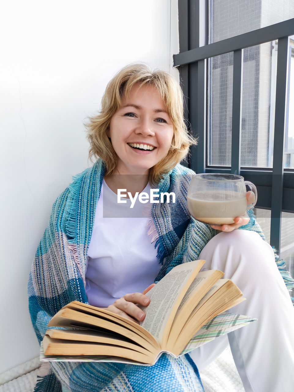 Portrait of a smiling young woman with short blond hair holding a book and coffee cup