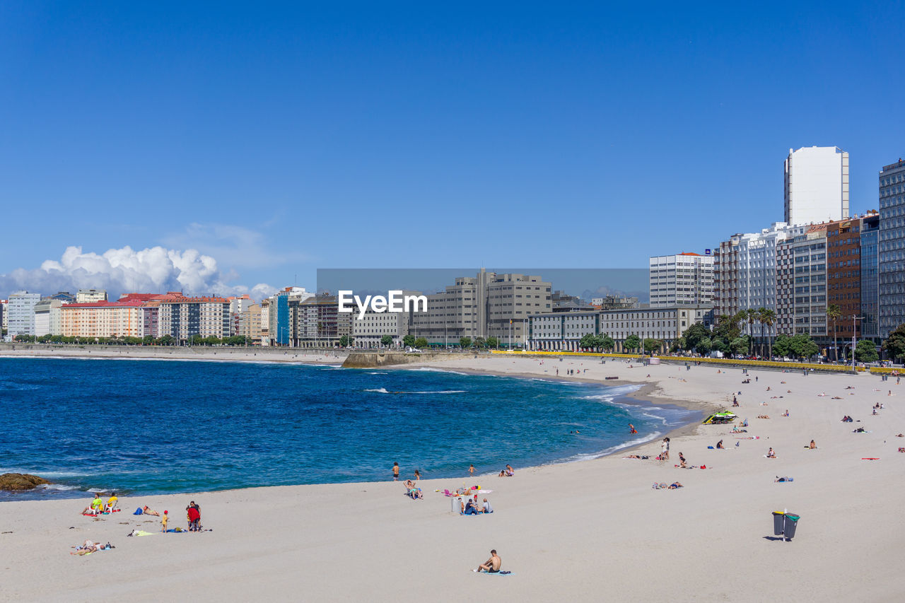 People on beach by buildings against blue sky