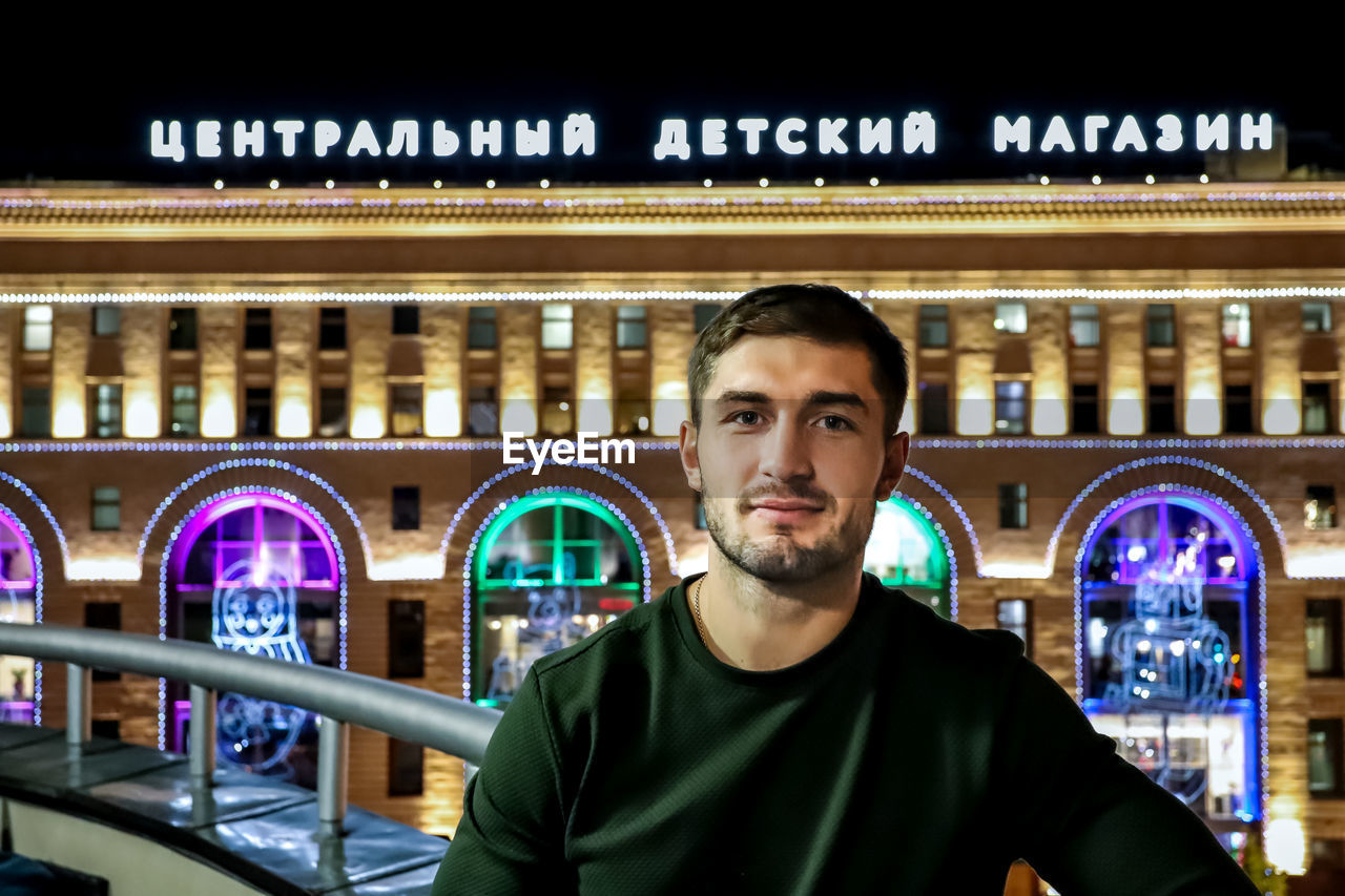 PORTRAIT OF YOUNG MAN STANDING AGAINST ILLUMINATED WALL