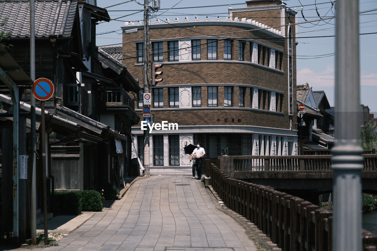 REAR VIEW OF MAN WALKING ON FOOTPATH AMIDST BUILDINGS IN CITY