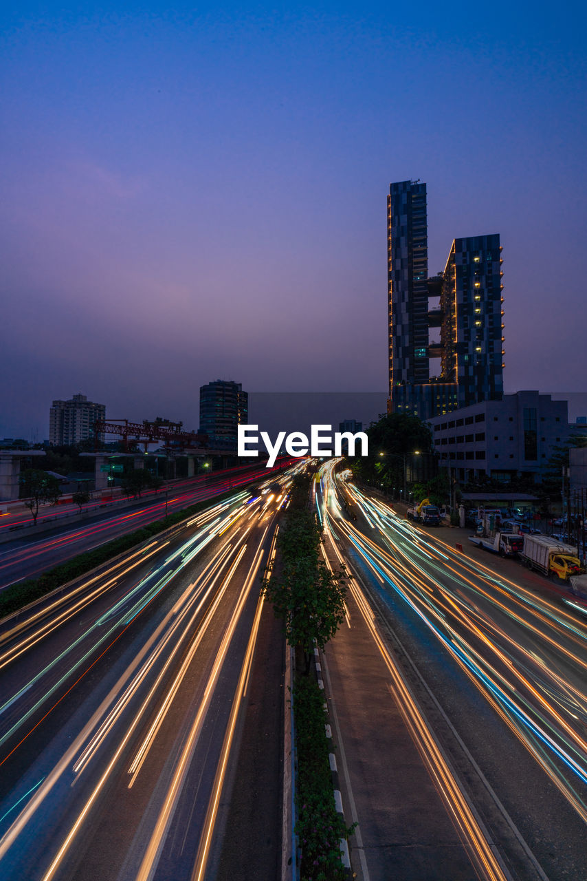 Light trails on road amidst buildings against sky at night