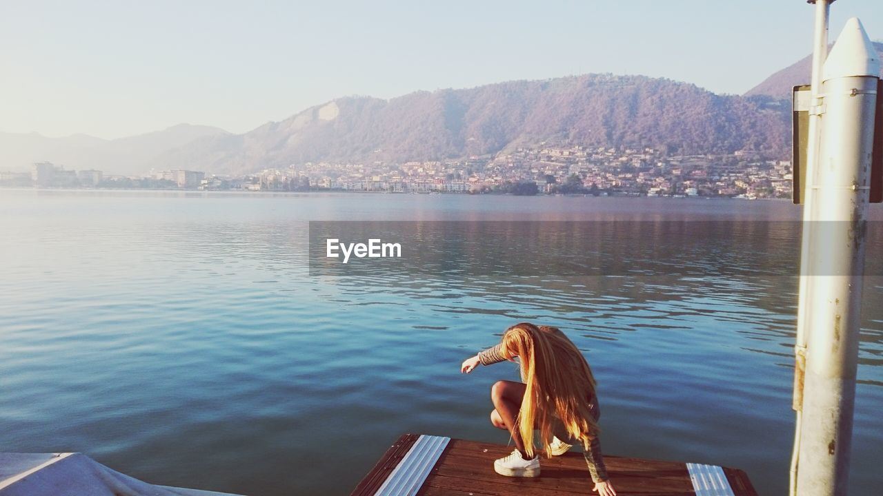 Woman crouching on pier over lake against sky