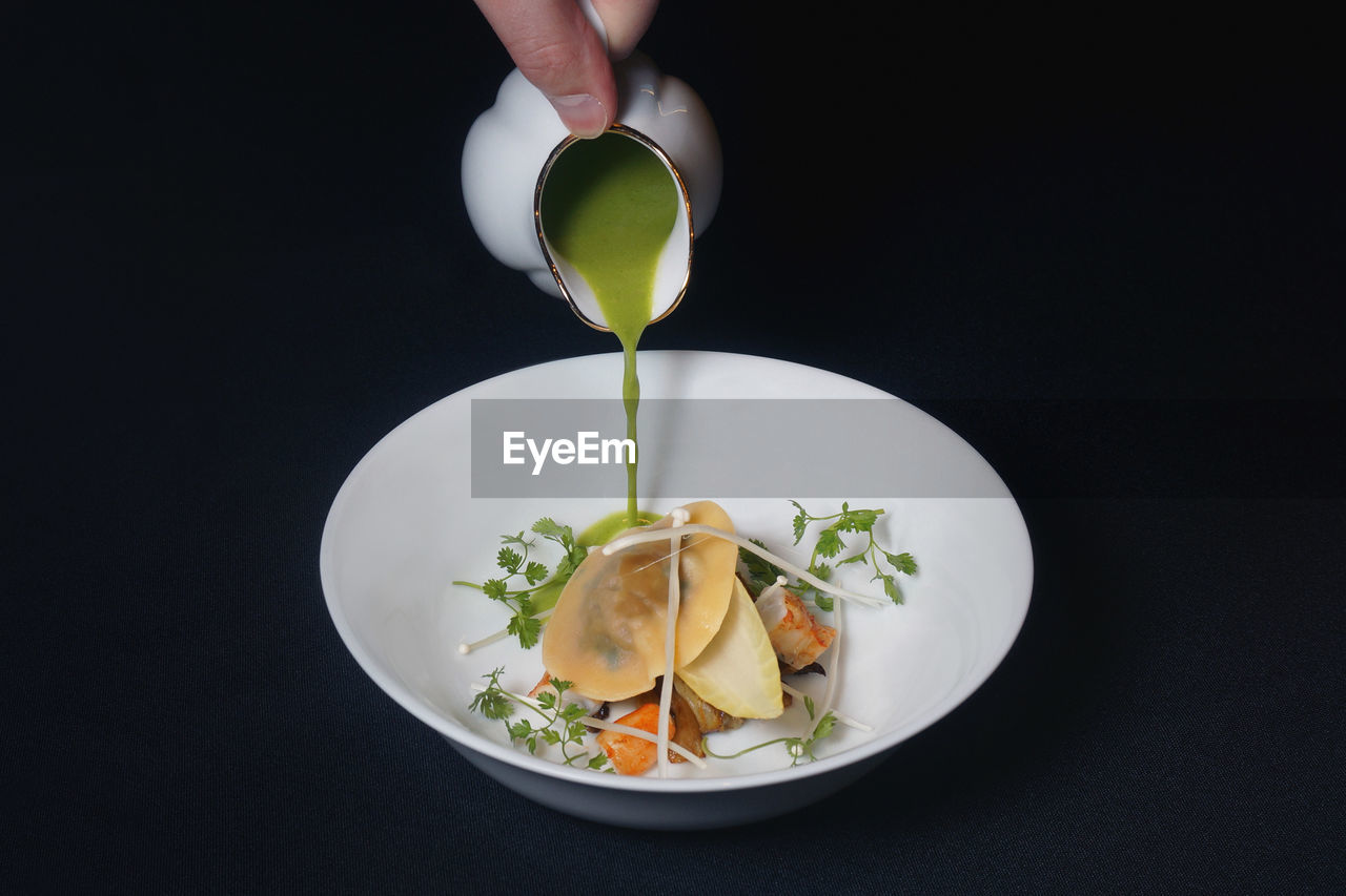 Close-up of hand pouring green liquid in food on bowl over black background