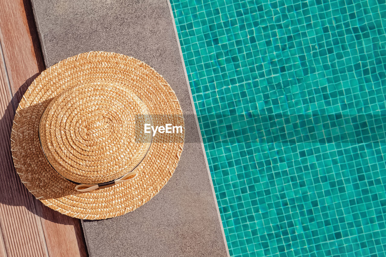 Woman's straw hat lying on the edge of the swimming pool with turquoise water, top view. 