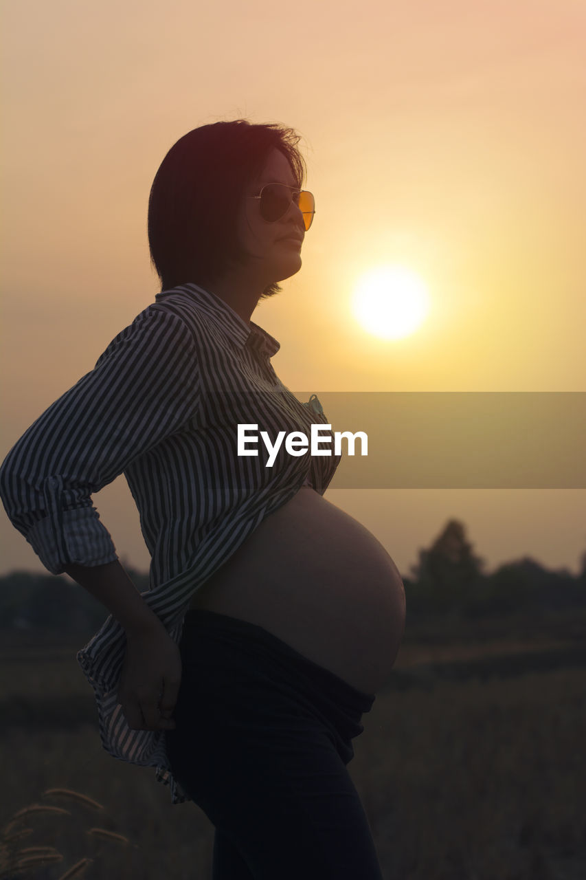 Pregnant woman wearing sunglasses standing against sky during sunset
