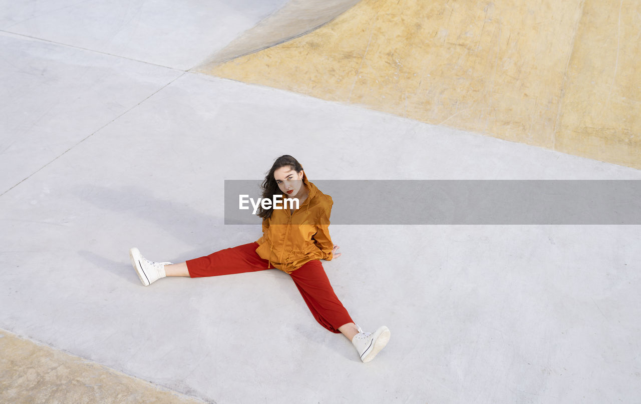 From above full body young female in stylish outfit sitting in concrete skate park while looking at camera in sunlight