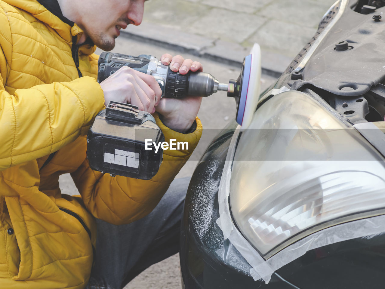 A  guy in a yellow jacket holds a drill with a microfiber disc and polishes the front headlight