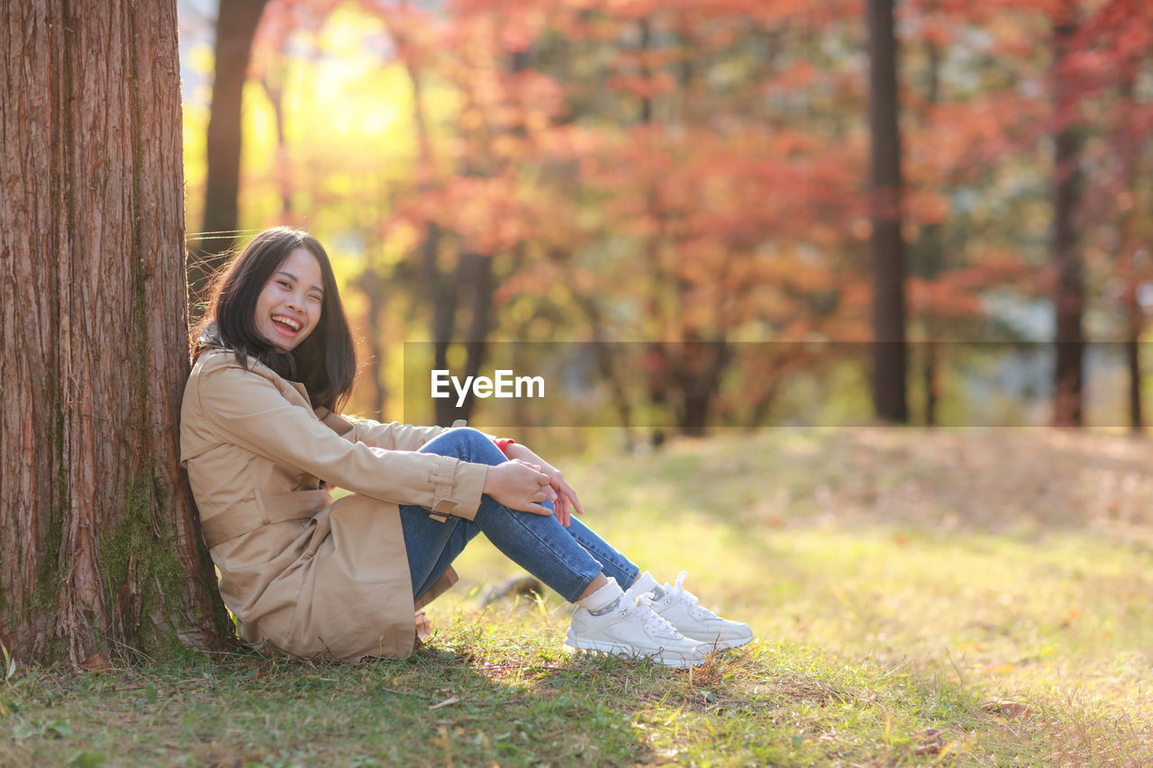 PORTRAIT OF SMILING YOUNG WOMAN SITTING ON TREE TRUNK DURING AUTUMN