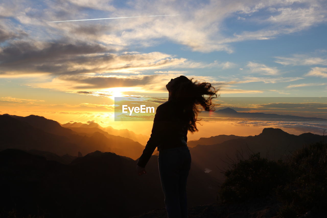 Silhouette young woman standing on mountain against sky during sunset