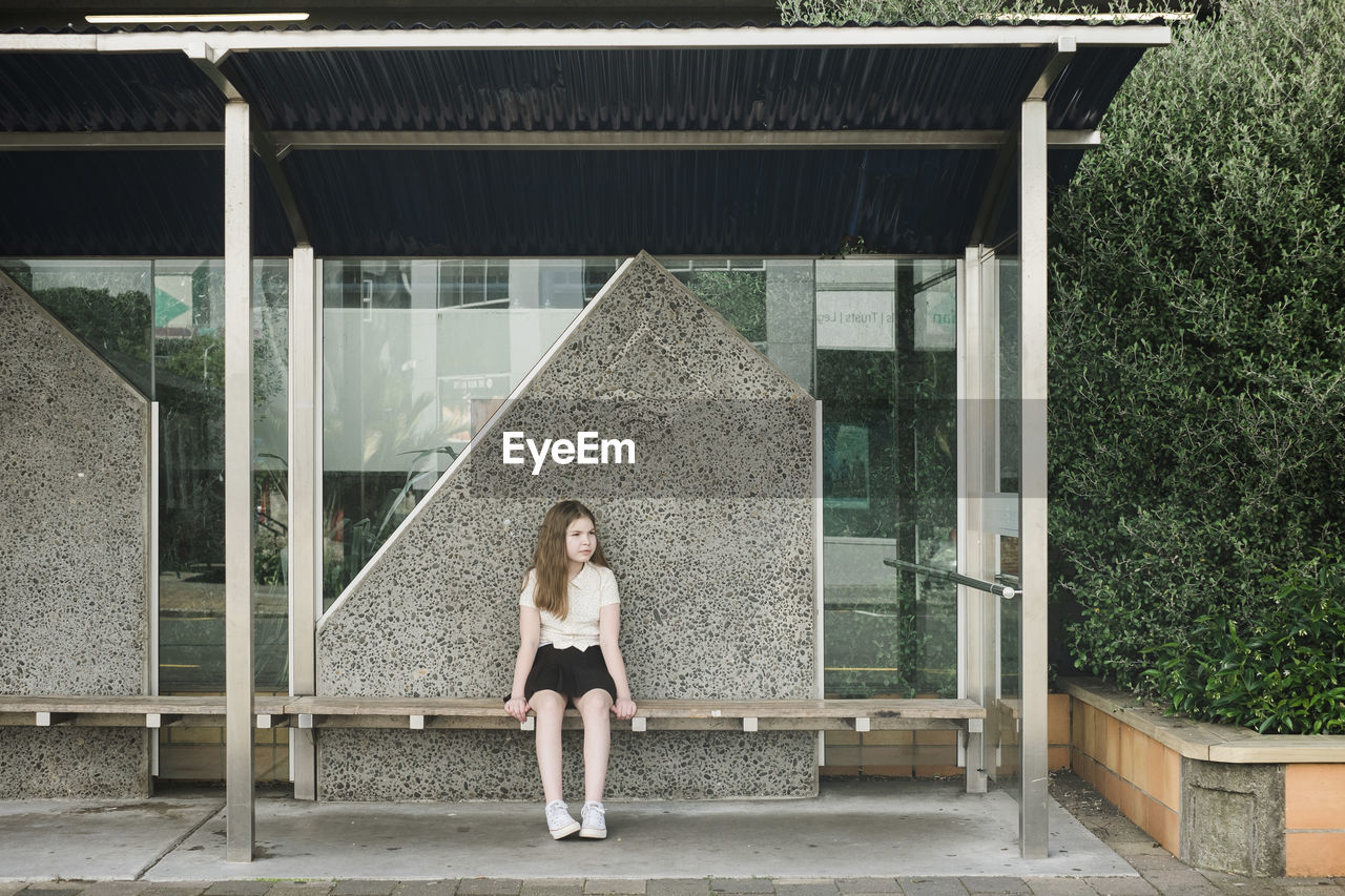 Young girl sitting alone on a wooden seat at an empty bus stop