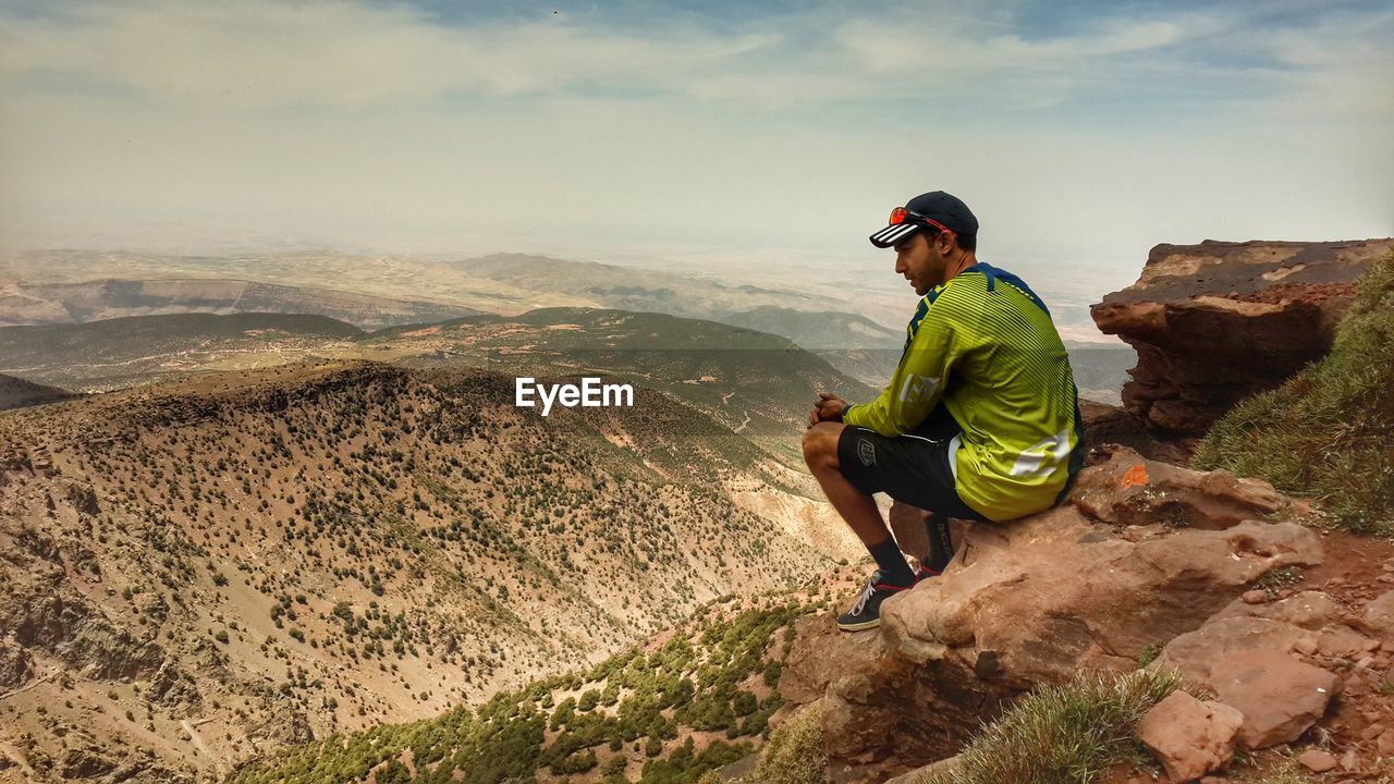 MAN SITTING ON MOUNTAIN AGAINST SKY