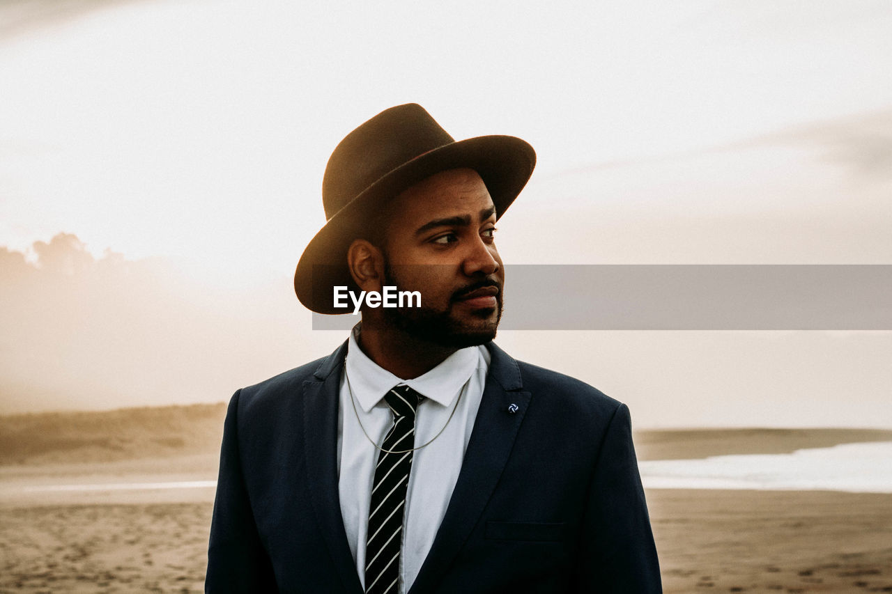 Young man in suit looking away while standing at beach against sky