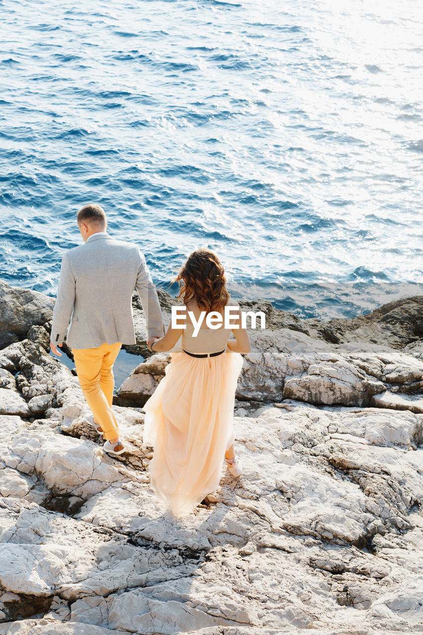 REAR VIEW OF COUPLE WALKING ON ROCK AT SEA SHORE