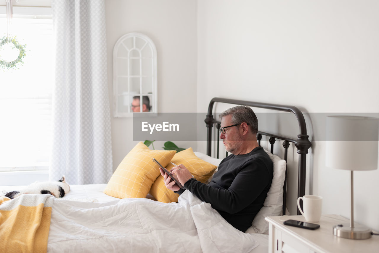 Man working on a digital tablet while sitting in bed in a light and bright bedroom