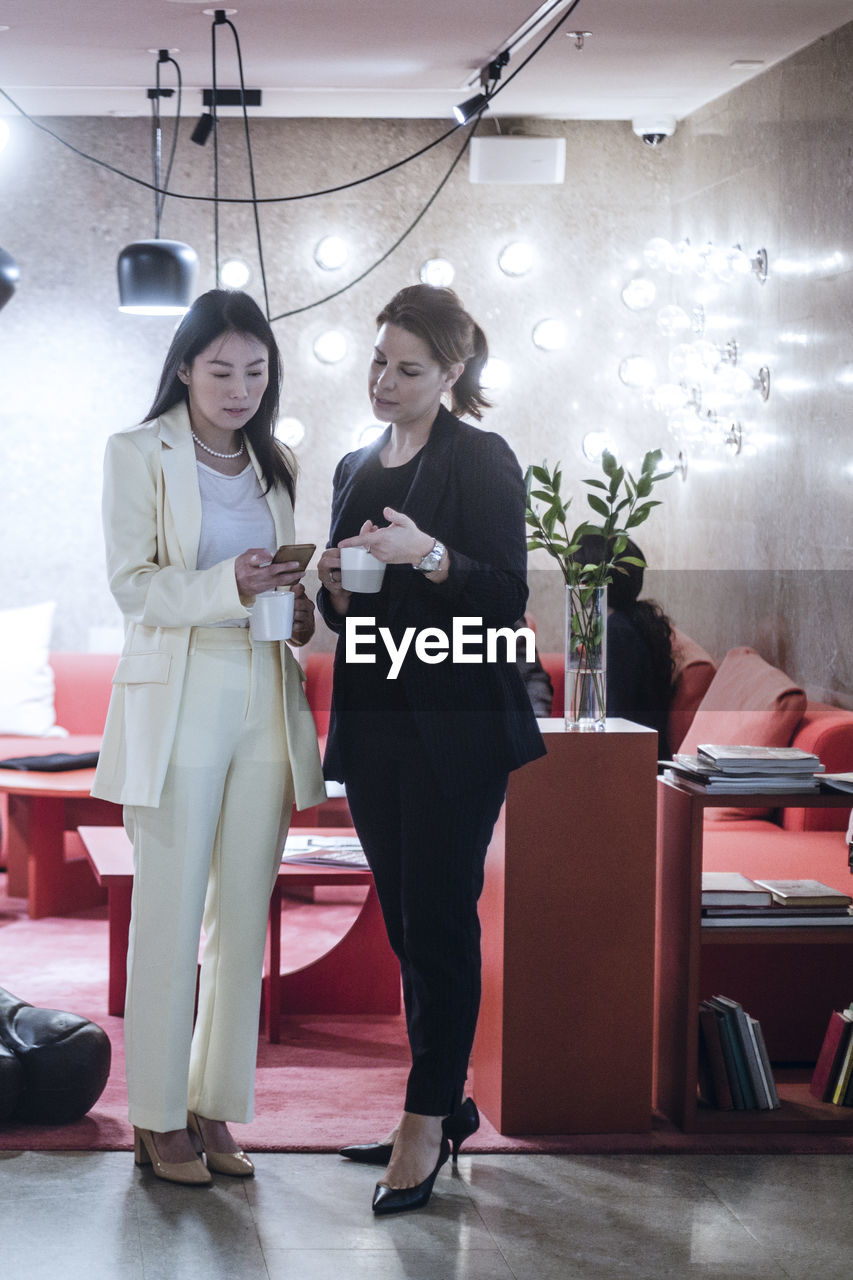 Full length of female coworkers talking during coffee break while colleagues working in background