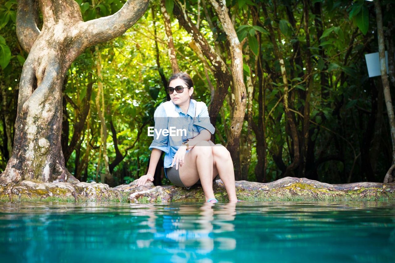 Young woman sitting by turquoise pond against trees