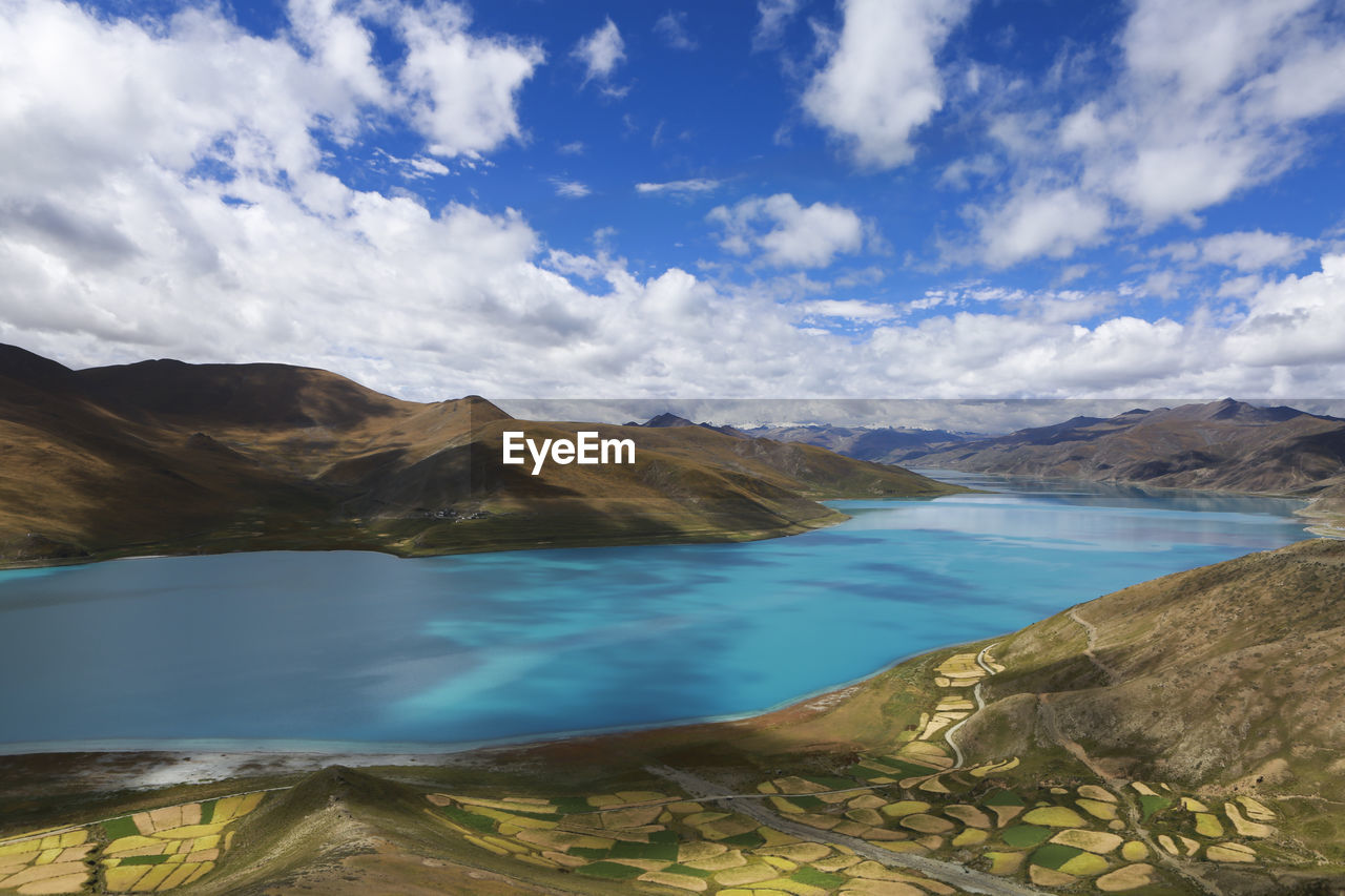 View of patched land with mountains and lake against cloudy sky