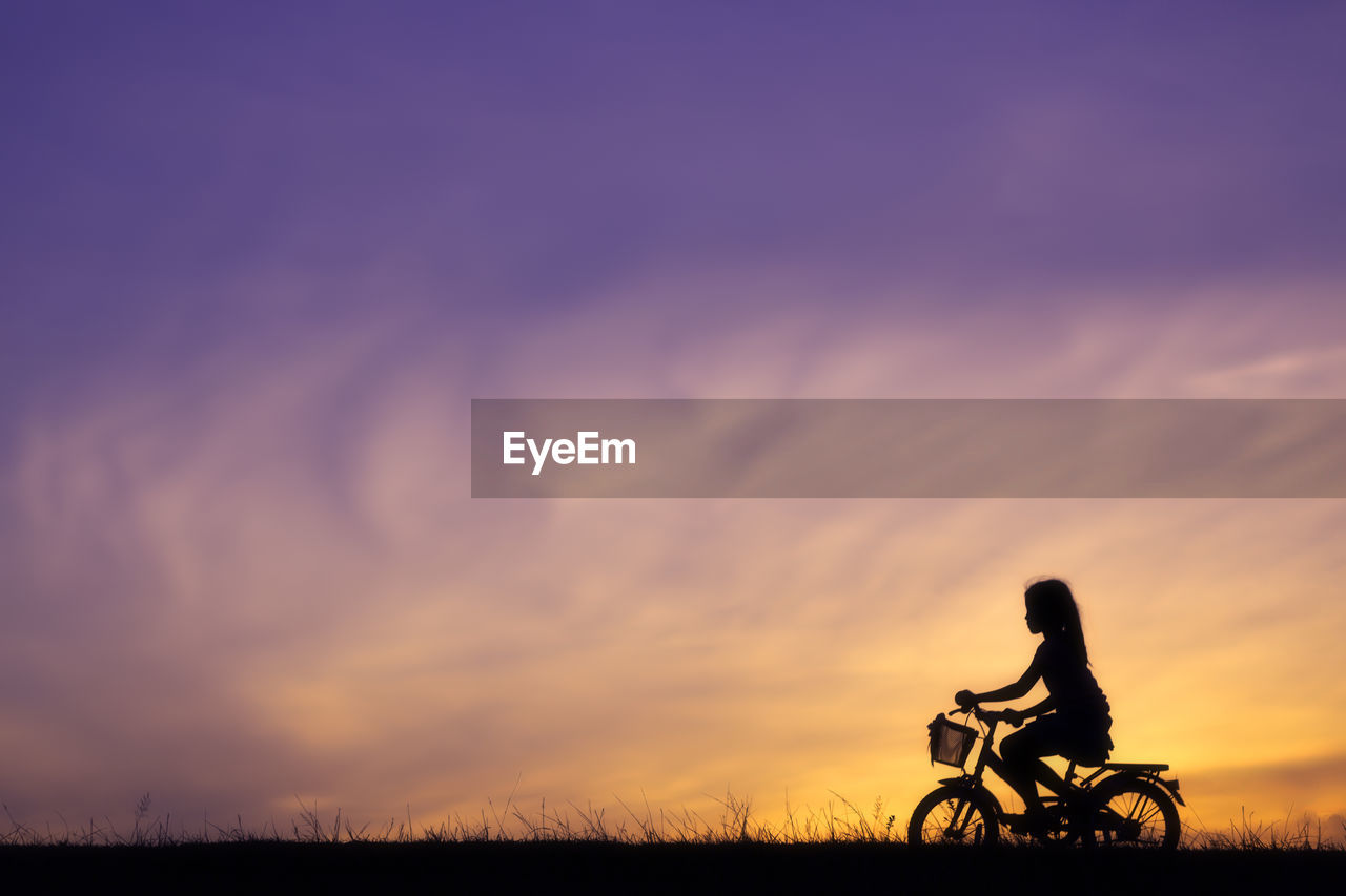 Silhouette girl riding bicycle against sky during sunset