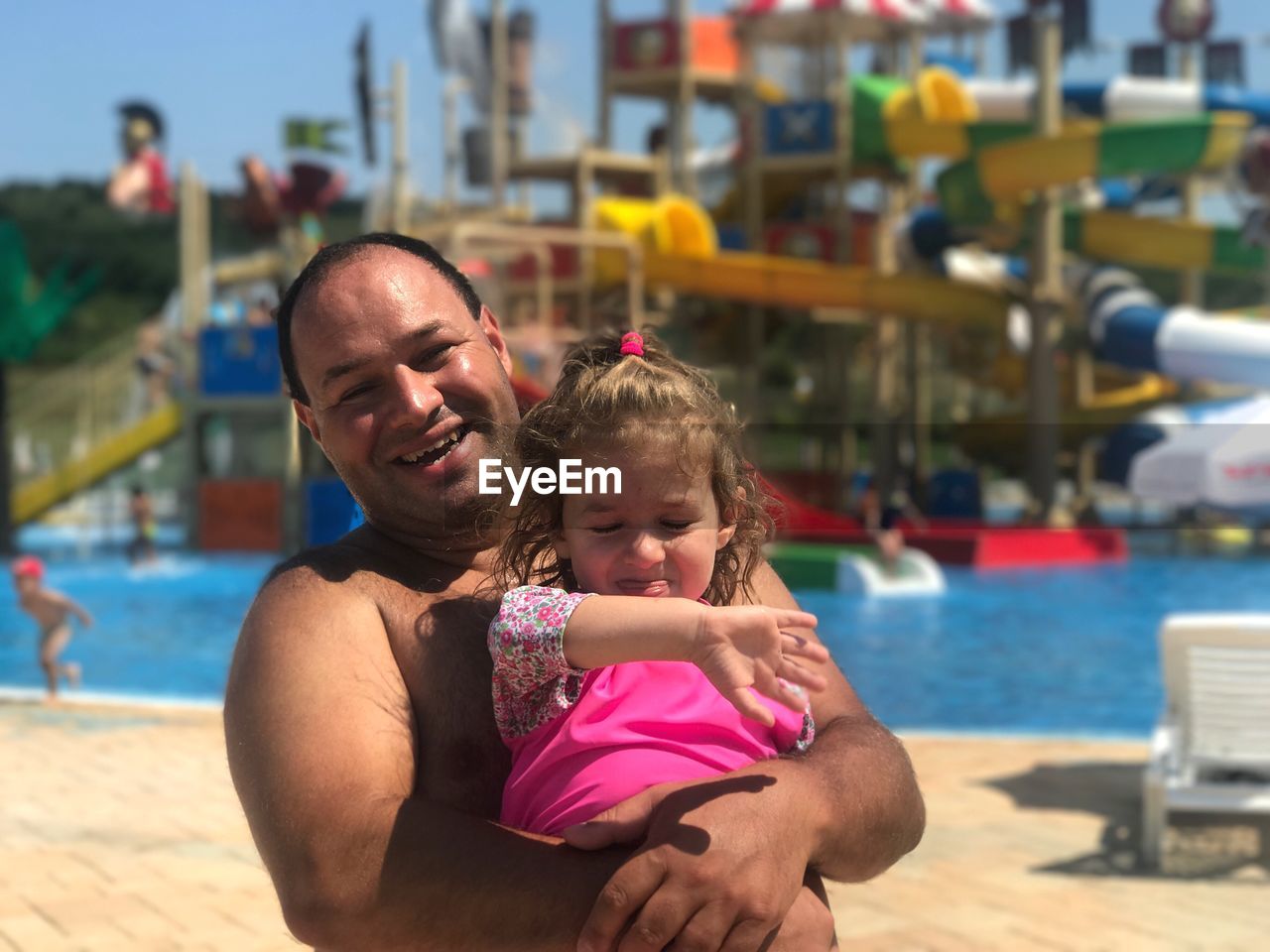 Portrait of happy shirtless father carrying daughter while standing at water park