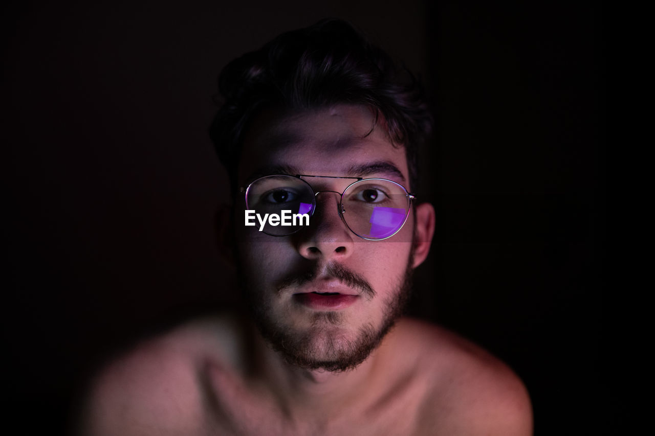 Close-up portrait of shirtless young man wearing eyeglasses against wall