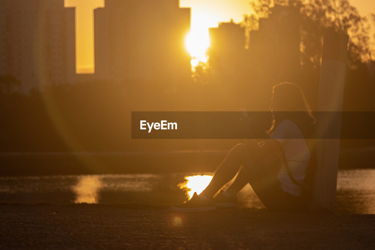 Side view of woman sitting at lakeshore during sunset