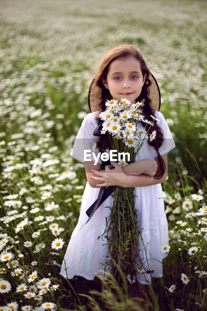 Portrait girl child in a white dress stands on a camomile field in a hat. bouquet of flowers