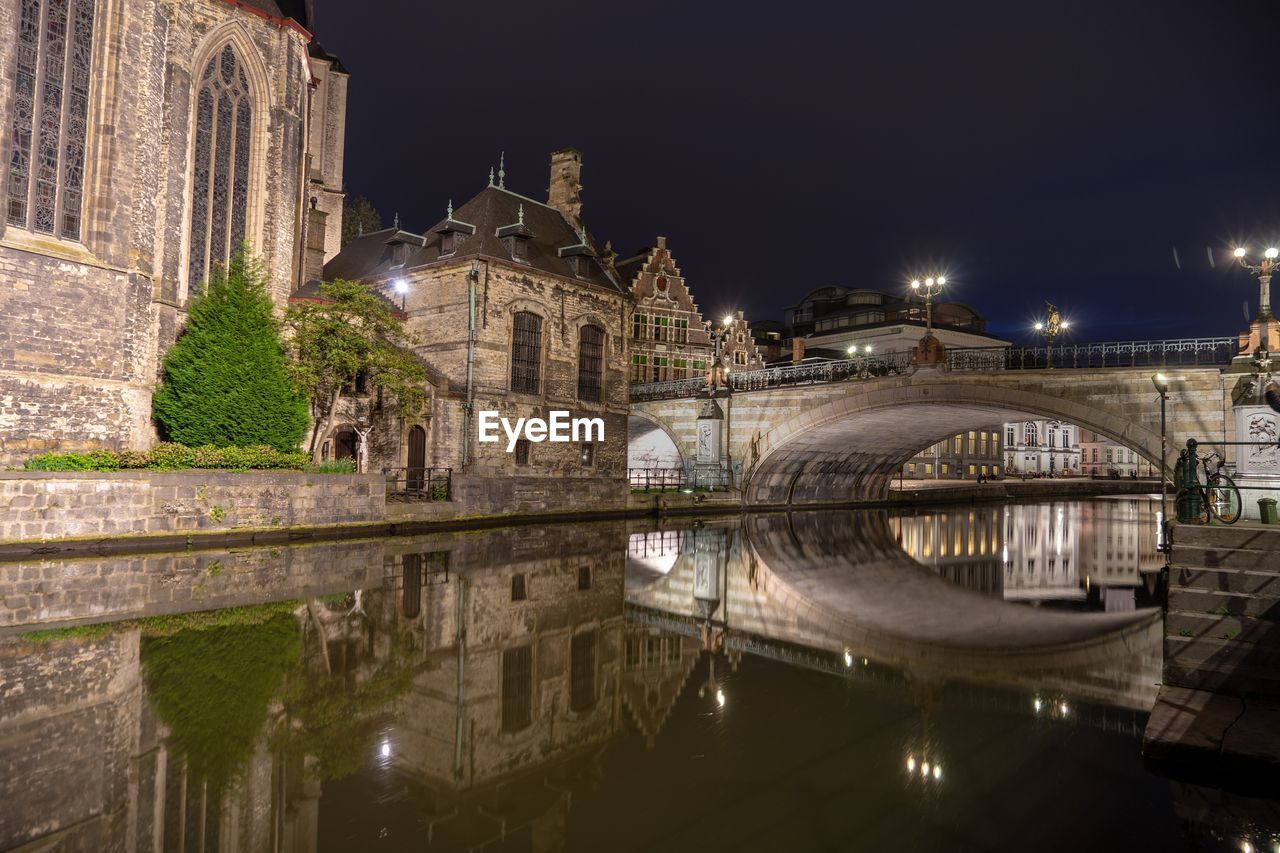 Illuminated bridge over river by buildings against sky at night