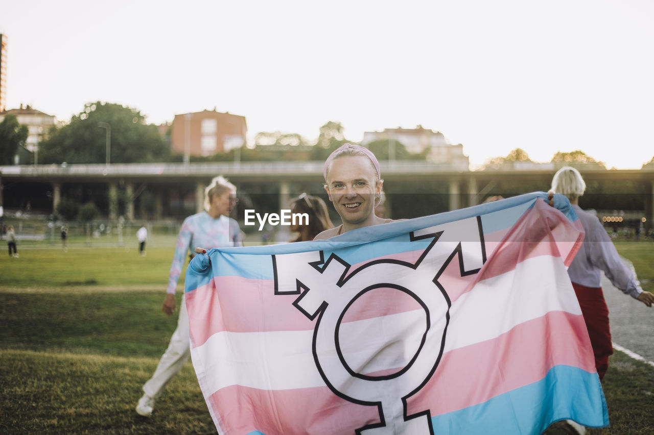 Portrait of happy non-binary person standing with transgender symbol on flag at park